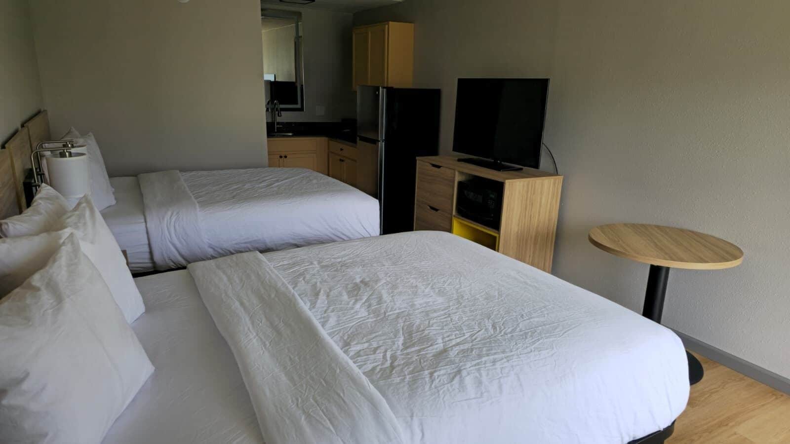 A hotel room with two beds, a TV, a small table, and a kitchenette.