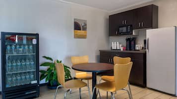 Cocina compacta con refrigerador, microondas, cafetera y dispensador de agua. Hay una pequeña mesa de comedor con cuatro sillas y una planta en maceta.