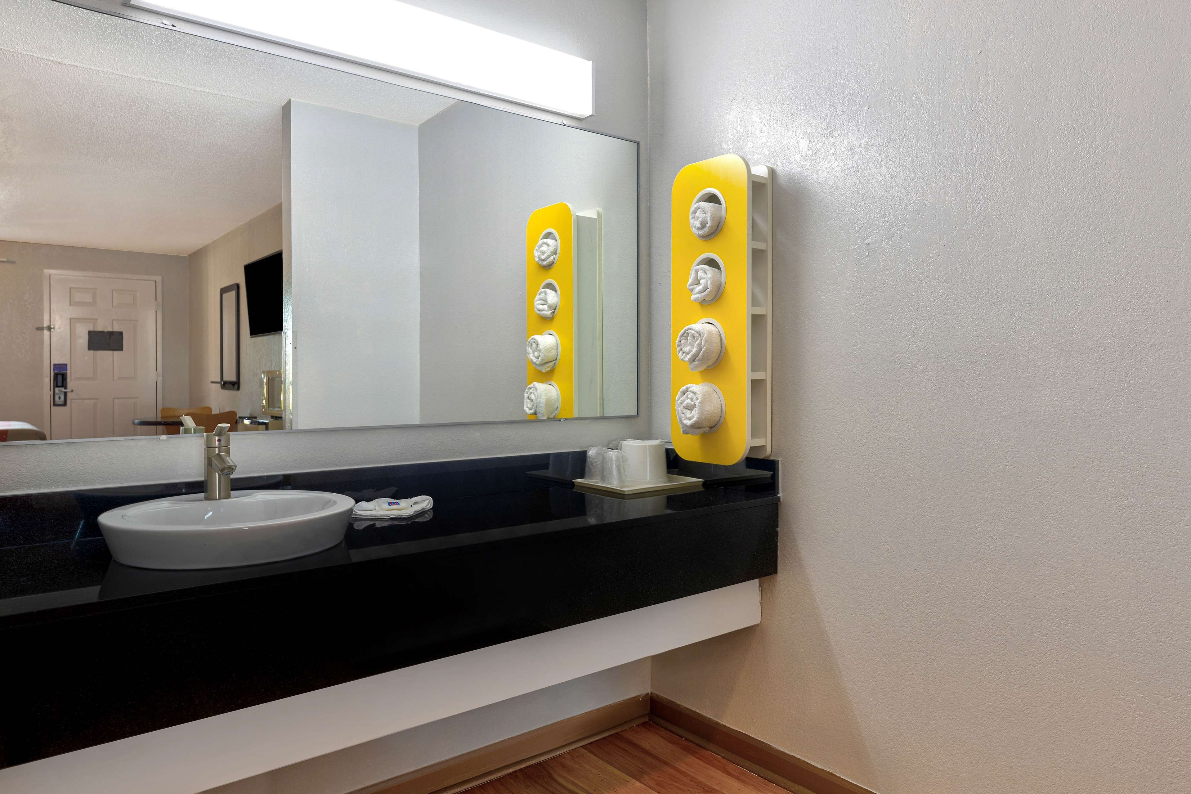 Een badkamer met een wit wastafel, een zwart aanrecht en een spiegel. Er zijn gele handdoekhouders en een witte beker op het aanrecht.
