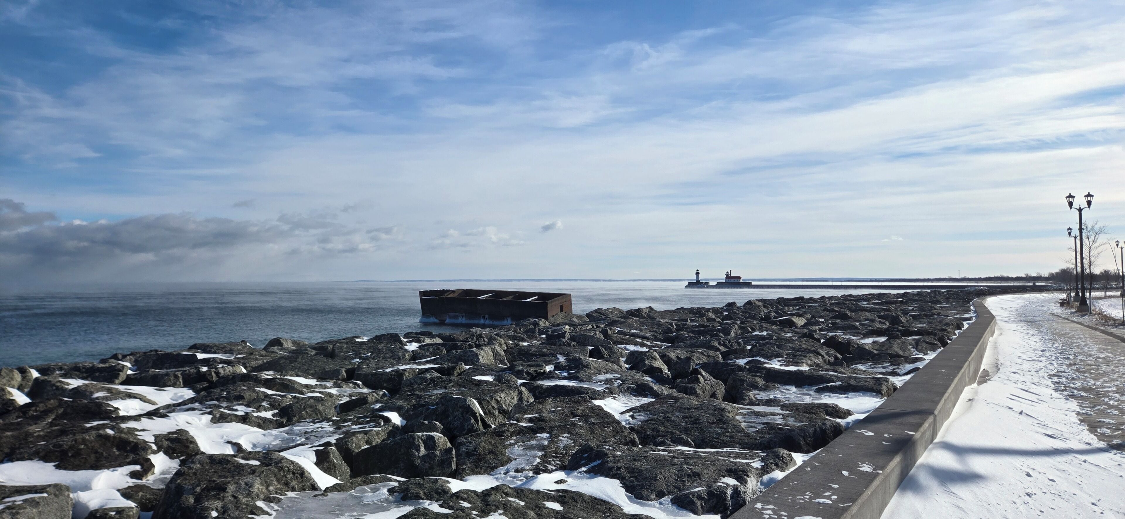 A rocky shoreline with a concrete walkway, a lighthouse in the distance, and a ship on the water.