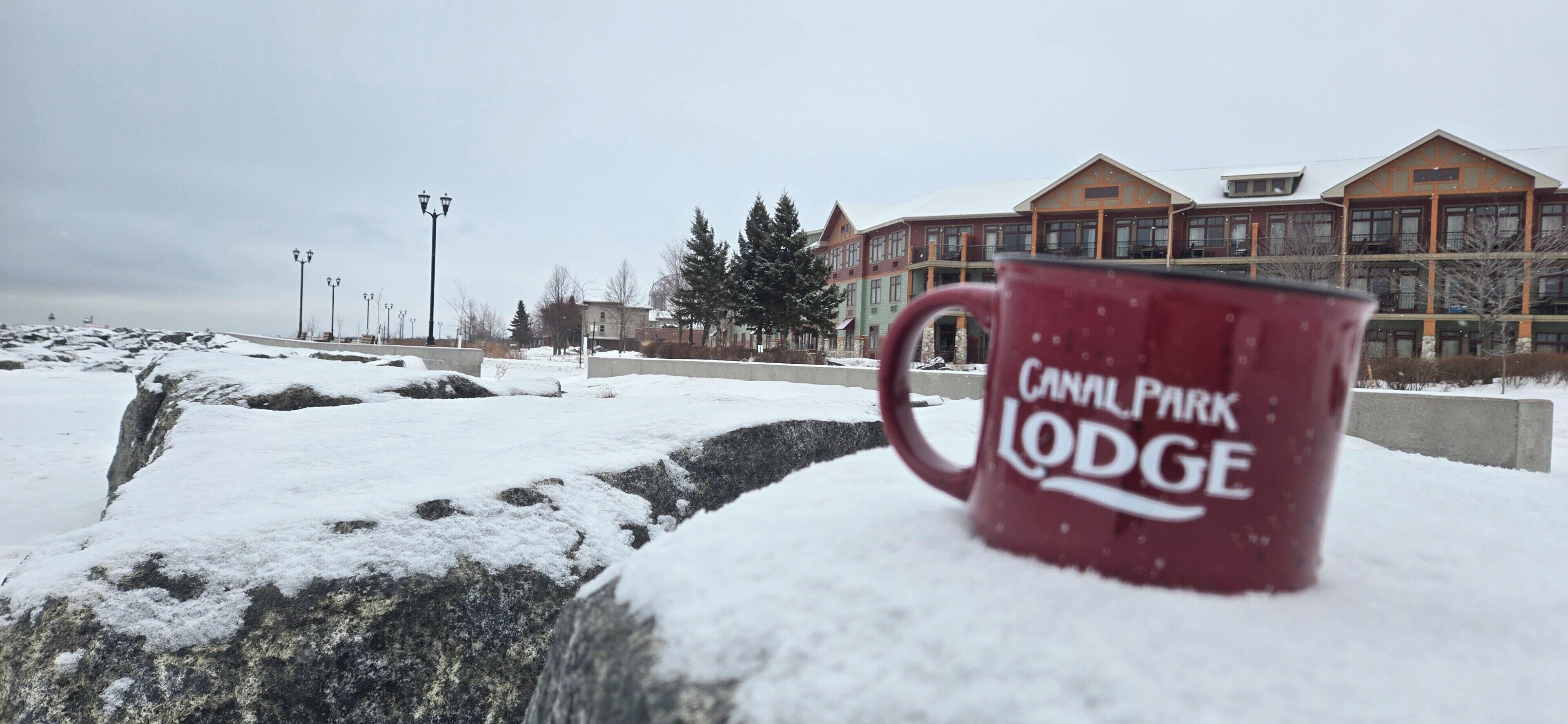 A snow-covered landscape with a red mug labeled 'CANAL PARK LODGE' in the foreground.
