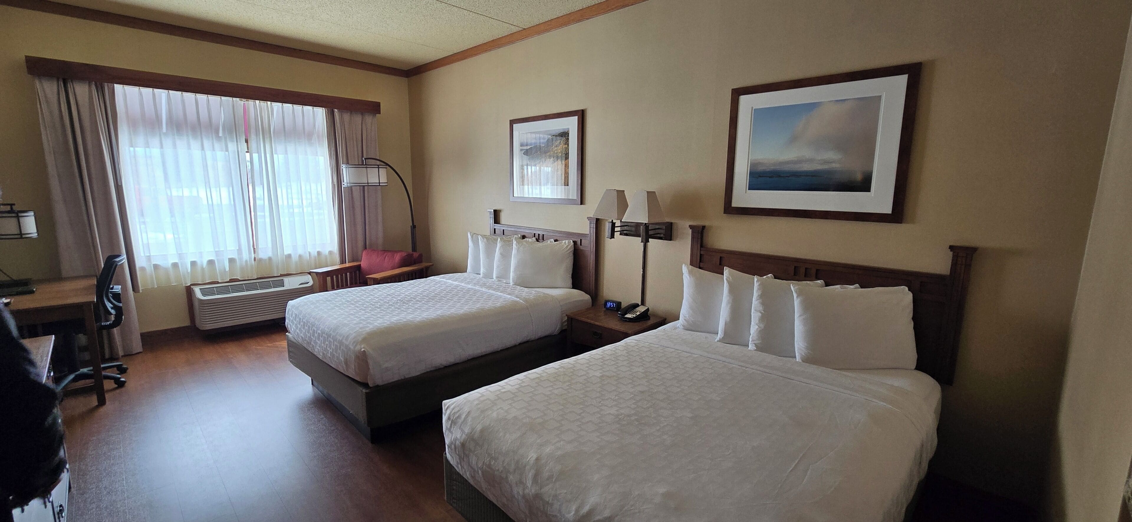A hotel room with two beds, a desk, a chair, a lamp, and a picture on the wall.