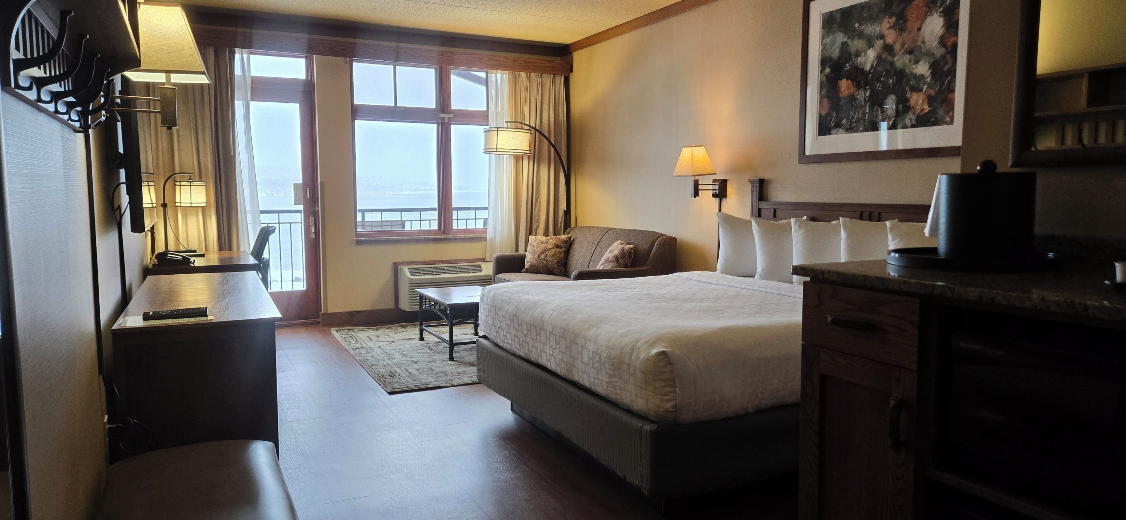 A hotel room with a bed, bedside table, lamp, desk, chair, and a view of the outside.