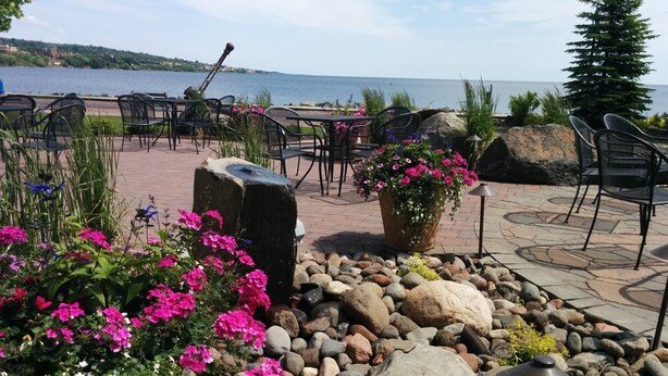 An outdoor seating area with tables and chairs, a rock garden with flowers, and a view of the water. - Canal Park Lodge (Duluth)