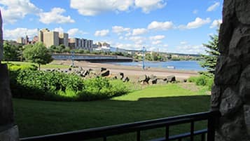 A view of a grassy area with a stone wall and a metal railing, overlooking a body of water and a distant cityscape.
