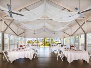 Un vaste espace intérieur aménagé avec des tables pour les repas, des ventilateurs de plafond et de grandes fenêtres offrant une vue sur les palmiers.
