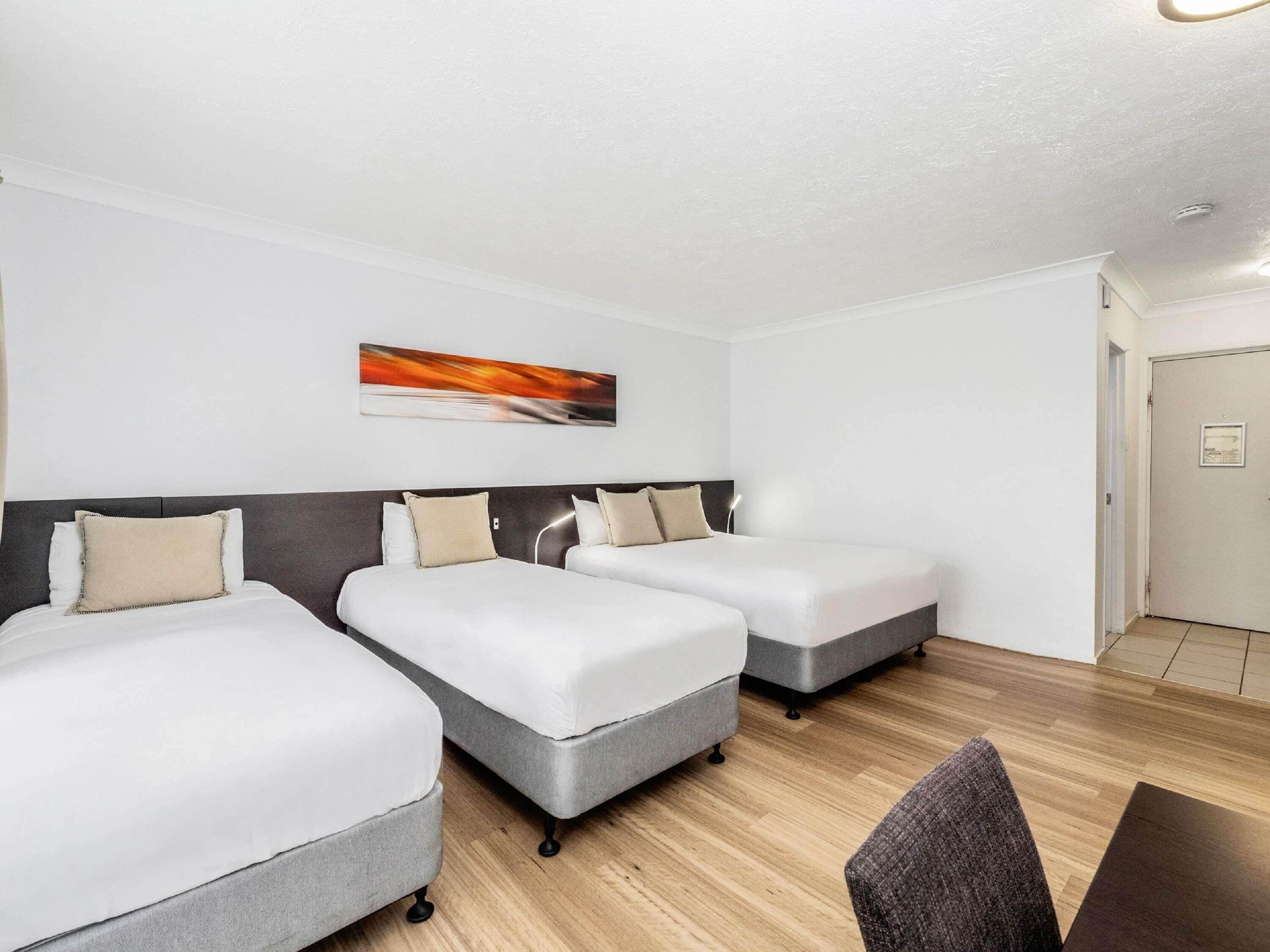 A hotel room with three beds, a wooden floor, a painting on the wall, and a door.