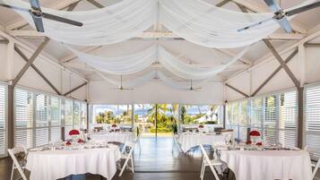 A spacious indoor area with tables set for dining, ceiling fans, and large windows offering a view of palm trees.