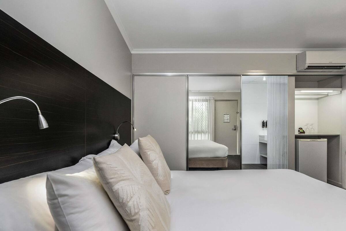 Une chambre d’hôtel moderne avec un grand lit, une lampe de chevet, un miroir et une petite kitchenette.