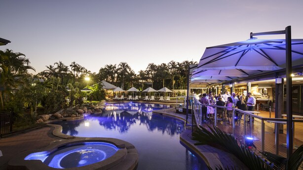 Zona de piscina con jacuzzi, sillones de descanso y sombrillas. Se observan palmeras y un edificio con zona de bar al fondo.