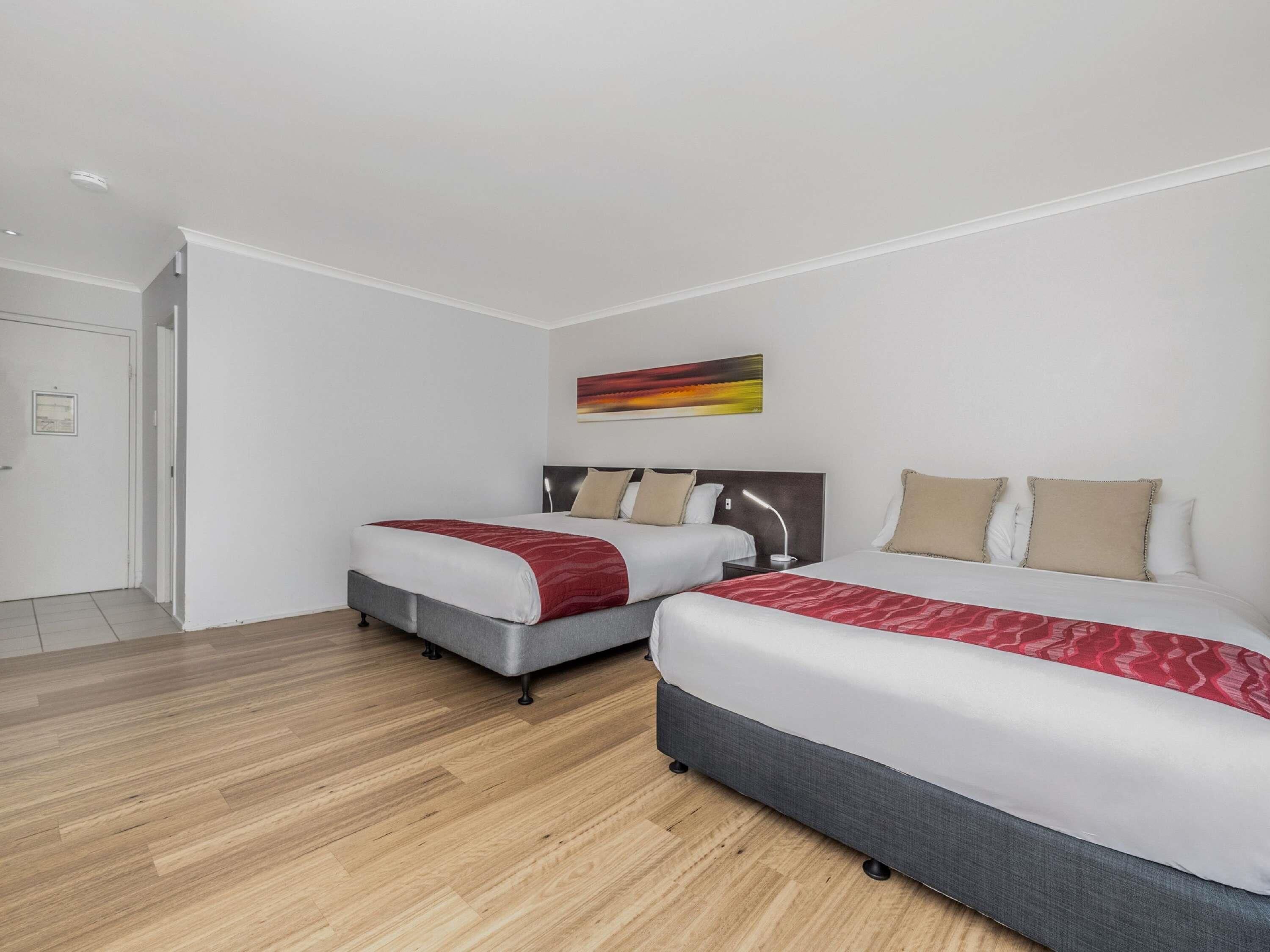 A hotel room with two beds, a wooden floor, a painting on the wall, and a door.