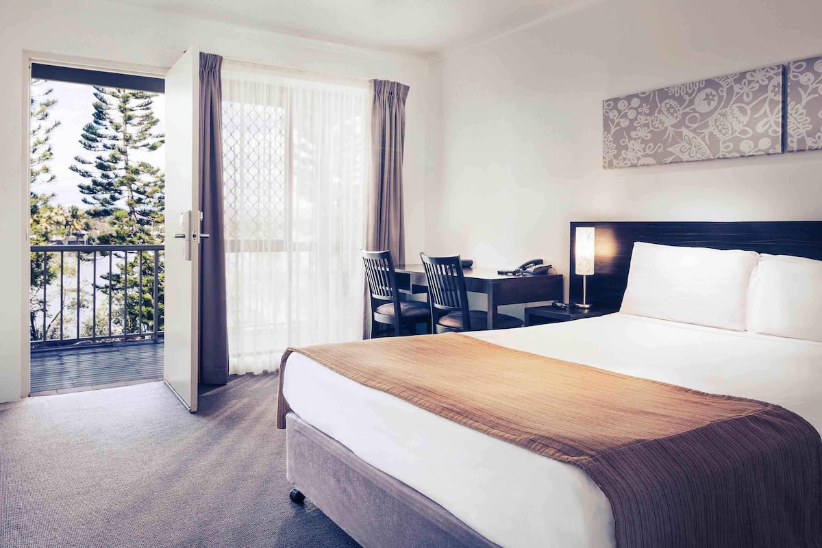 Habitación de hotel con cama, escritorio y silla. Tiene un balcón con vistas a árboles.