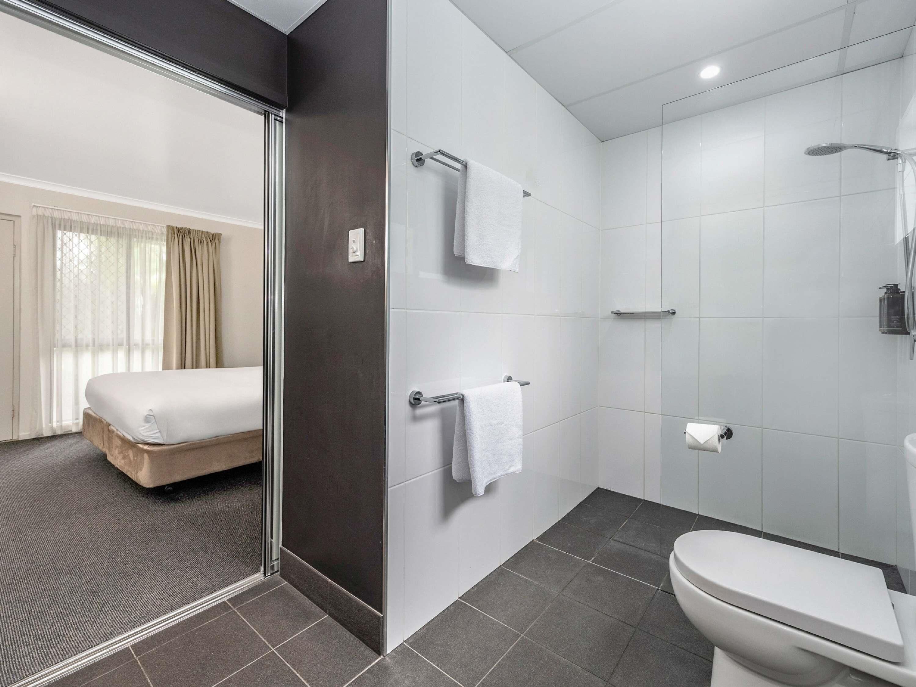 Casa de banho de hotel moderna, com chuveiro, sanita e uma cama visível através de uma porta de vidro.