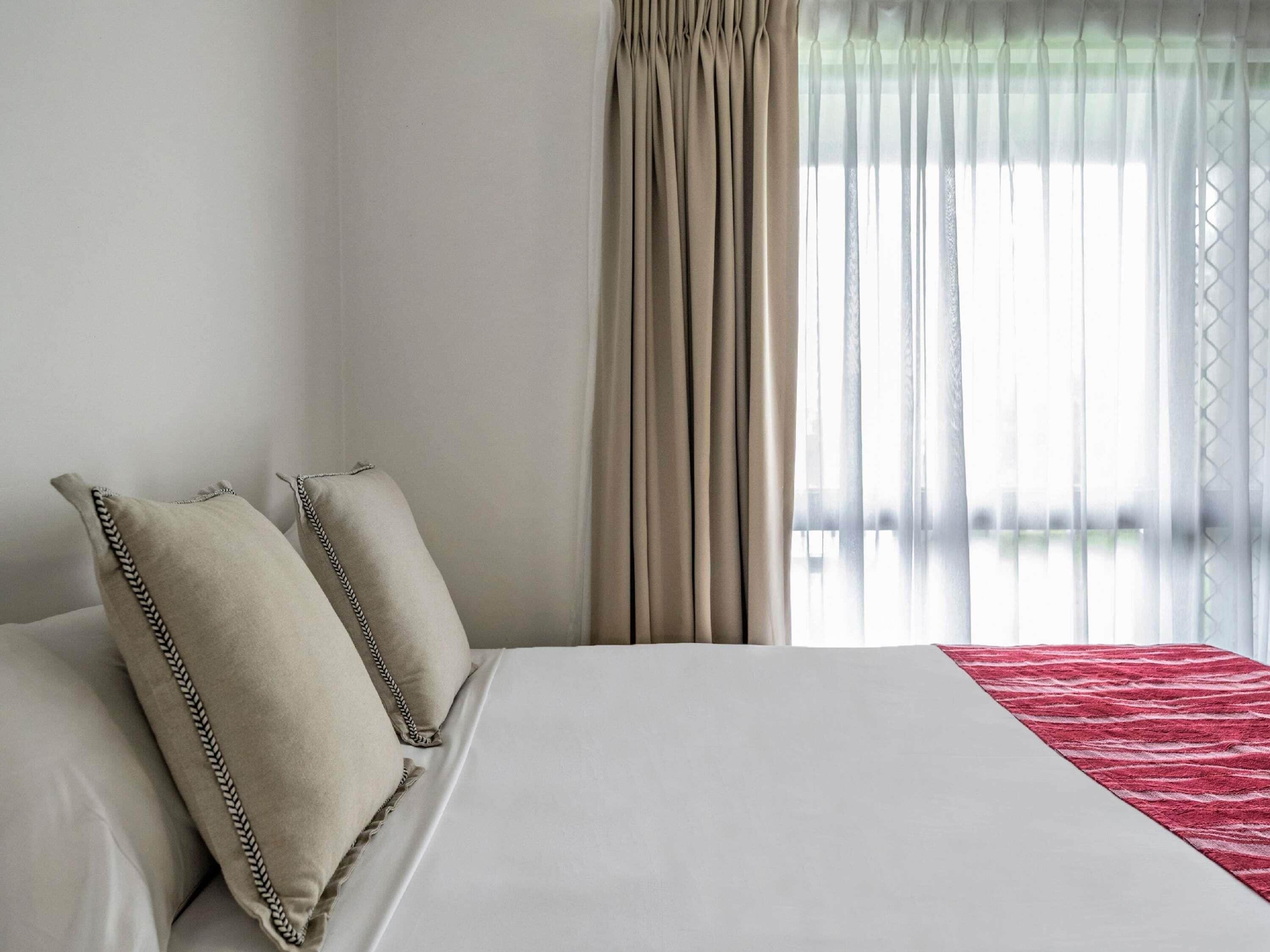 A neatly made bed with pillows, a red blanket, and sheer curtains.