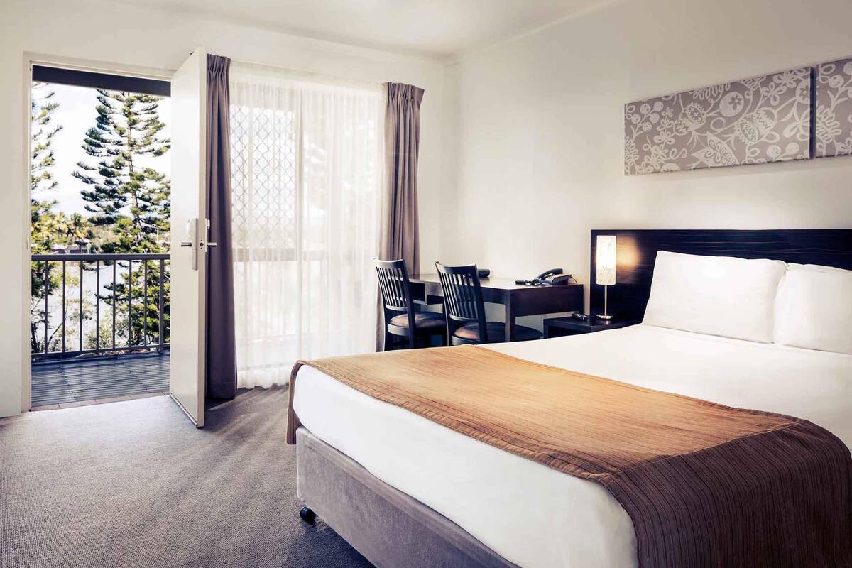 Une chambre d’hôtel avec un lit, un bureau, une chaise, un téléphone et une vue sur les arbres par la fenêtre.