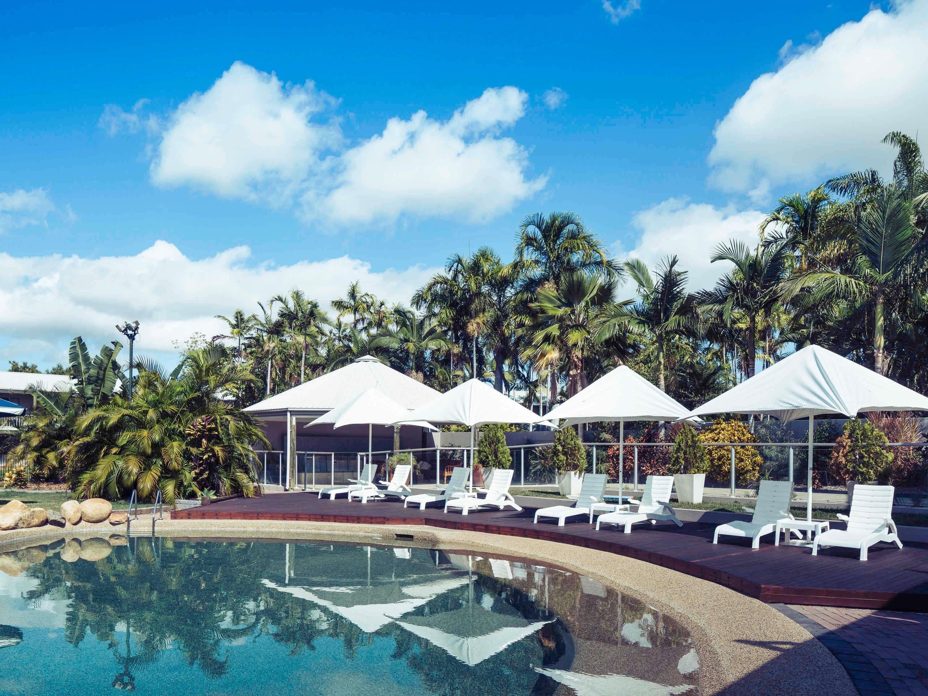 Un espace piscine agrémenté de chaises longues et de parasols, entouré de palmiers et surplombé d’un ciel dégagé.