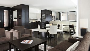 Een moderne woonkamer met donkere houten vloeren, een salontafel van donker hout en een bank. Er is een eethoek met witte stoelen en een tafel. De keuken heeft donkere kastjes en een wit aanrecht.