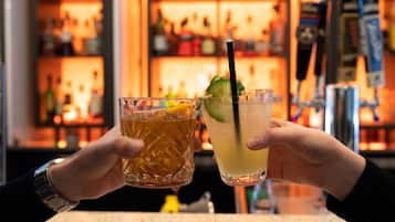 Dos manos sosteniendo vasos con bebidas, uno con una rodaja de limón y el otro con una rodaja de pepino, en un bar con estantes llenos de botellas al fondo.