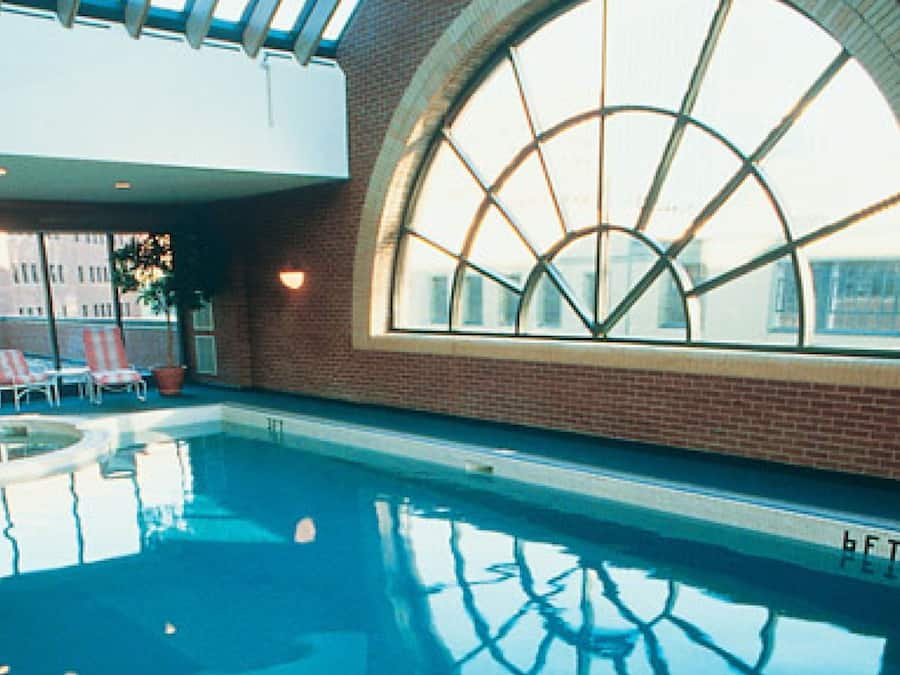 An indoor swimming pool with a large arched window, lounge chairs, and a brick wall.