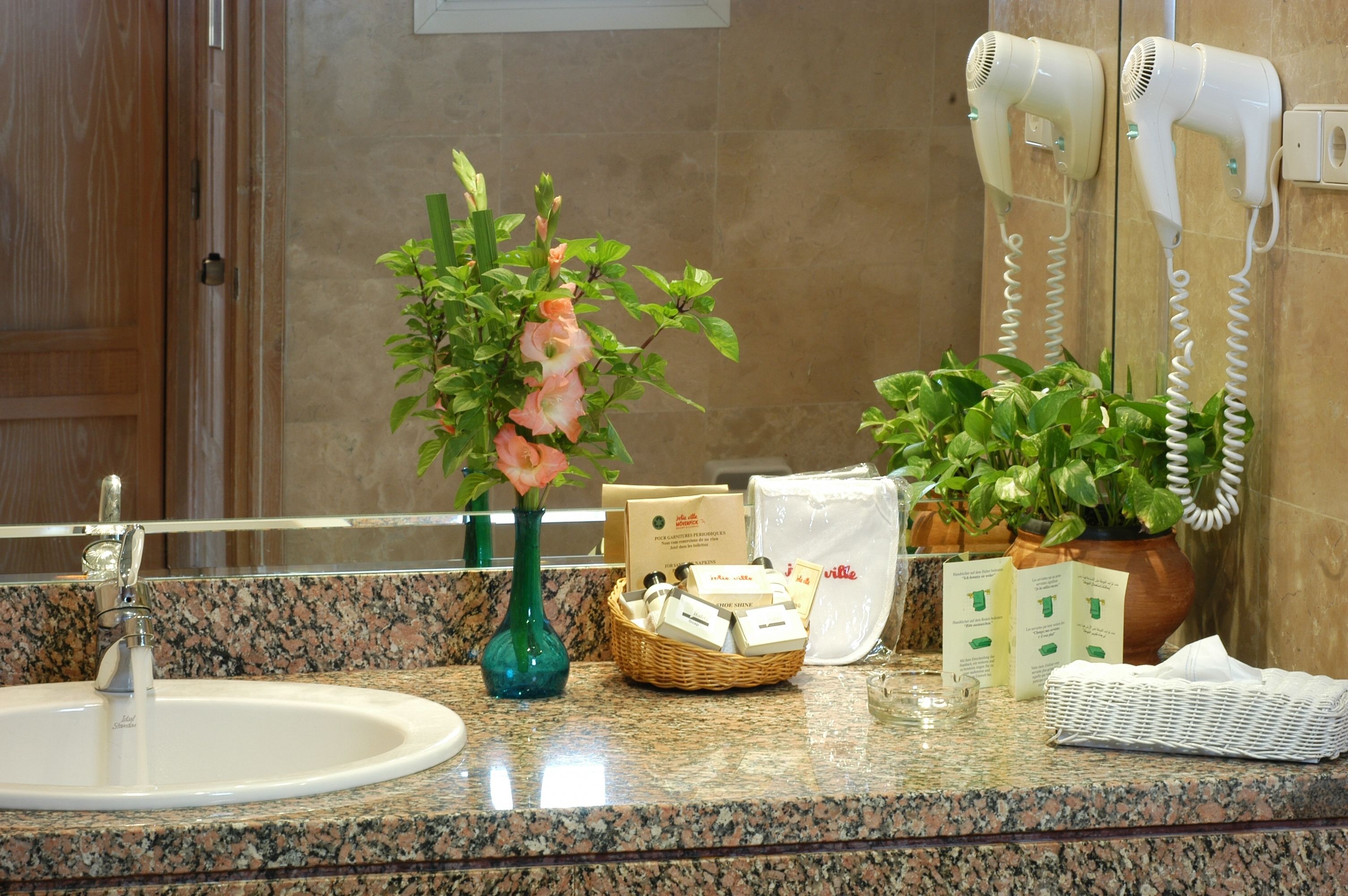 Ein Badezimmer mit einer Granittafel, einem weißen Waschbecken, einer grünen Vase mit Blumen, einem Fön und Pflegeprodukten.