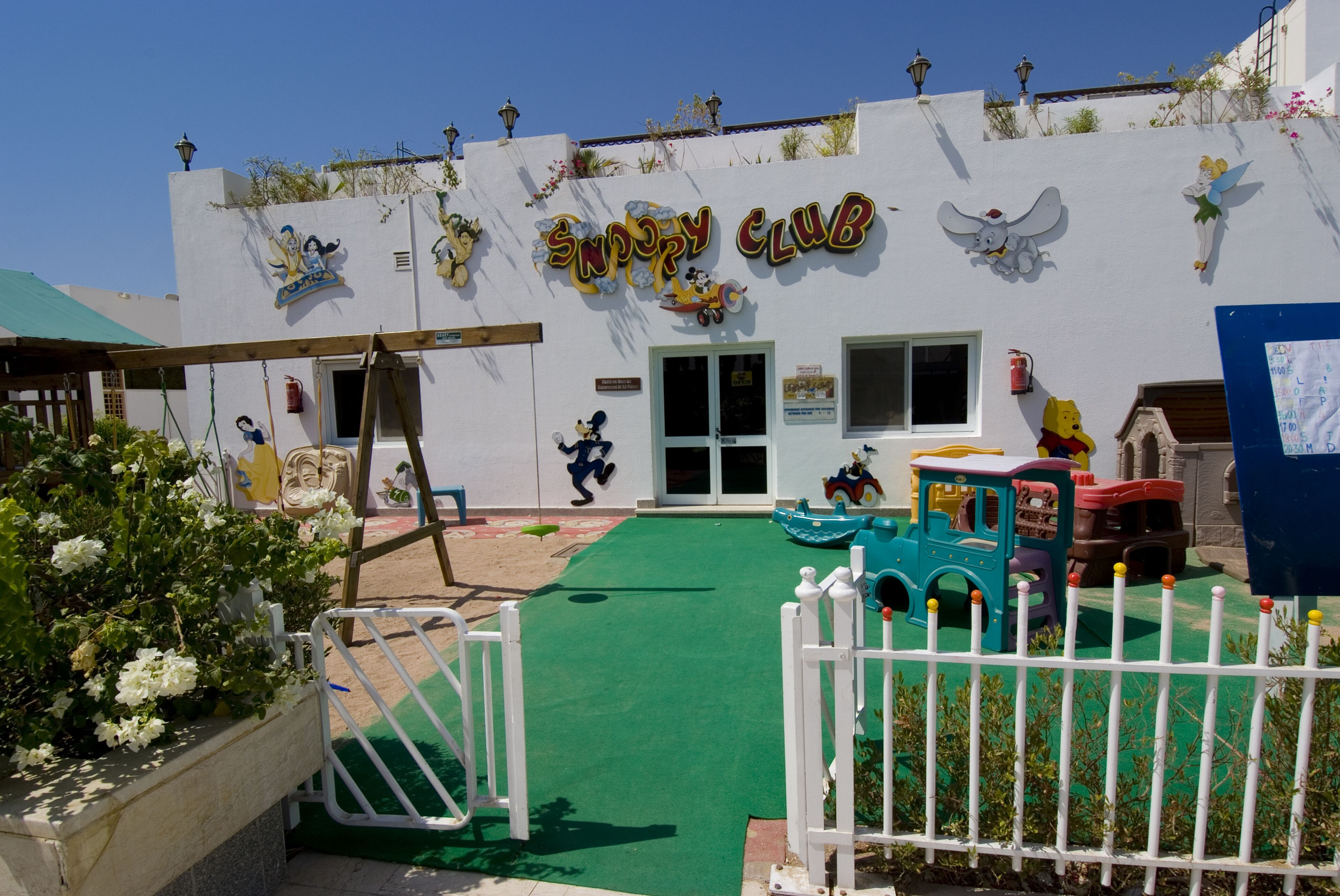 A building with a sign reading "Snoopy Club" and a play area with a green floor and a white fence.