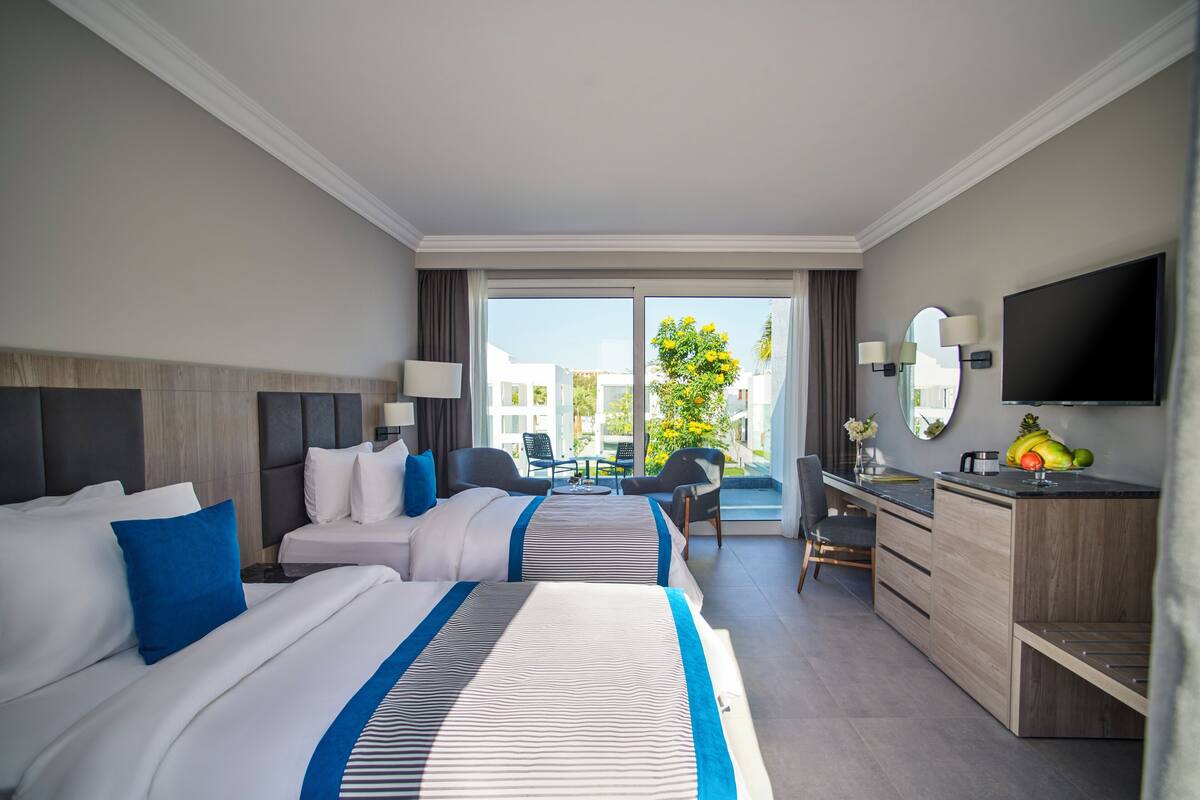 A hotel room with two beds, a desk, a TV, and a balcony with a view of buildings and trees.