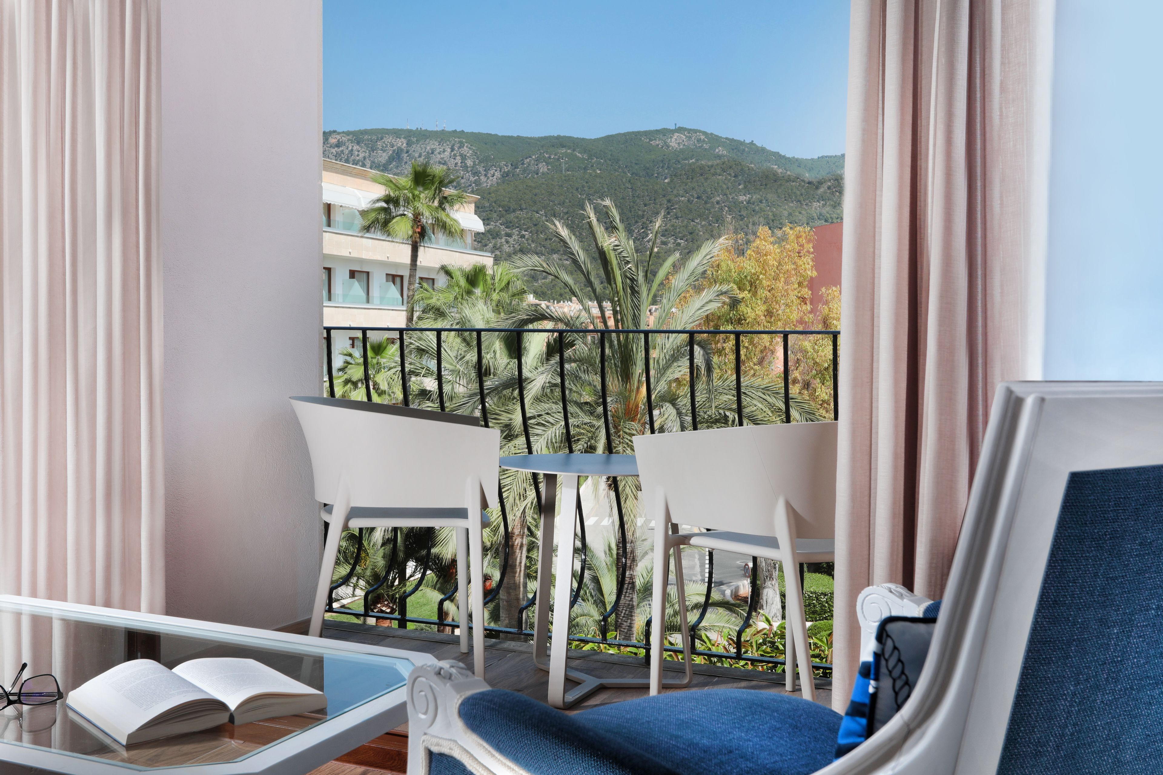 Un balcon avec une table en verre, deux chaises blanches, un canapé bleu et un livre ouvert.