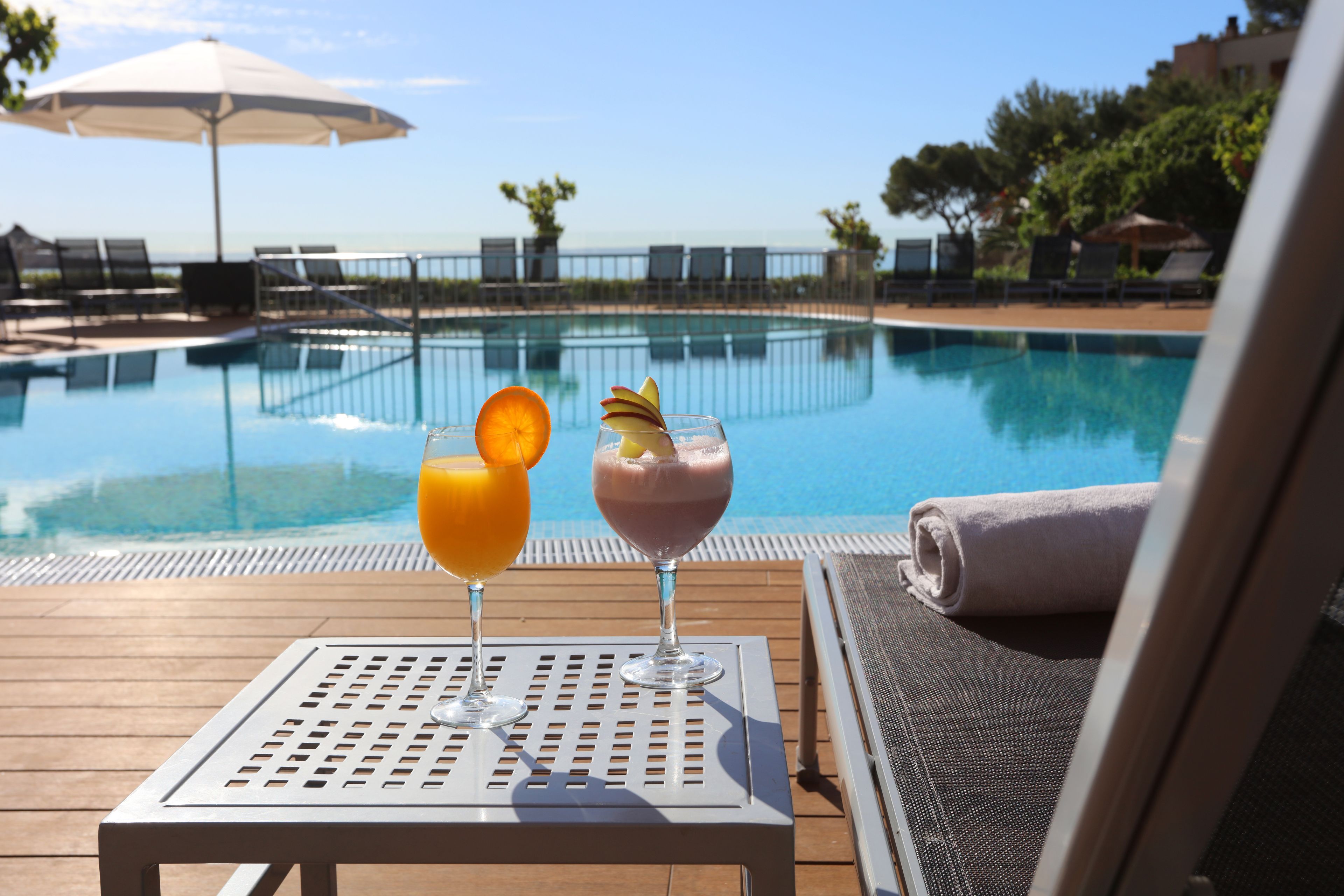 A poolside setting with two drinks on a table, a lounge chair, and a pool in the background.