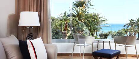 Habitación de hotel moderna con sofá, una mesa y sillas. Se ve una vista de palmeras y el mar a través de ventanas amplias.