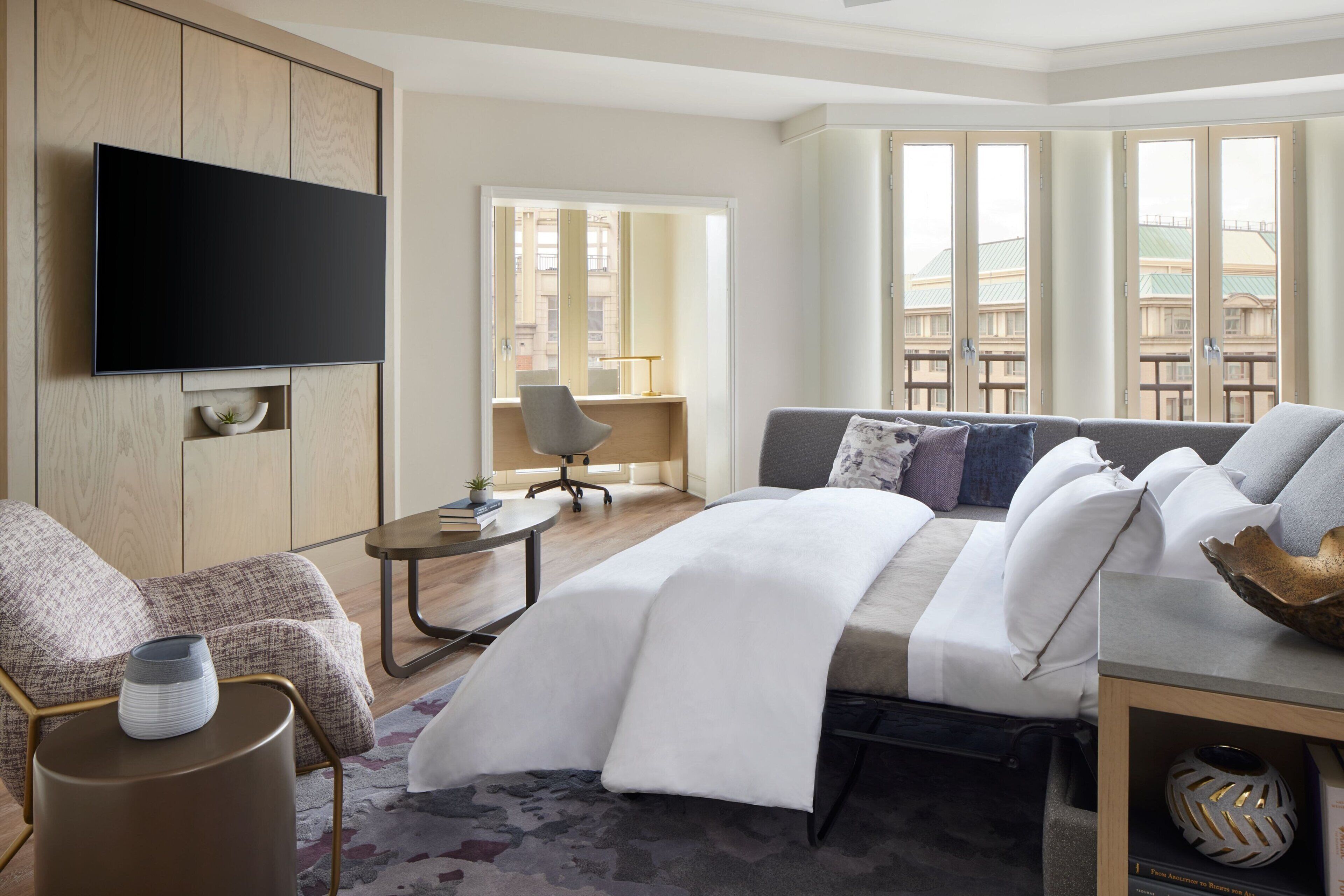 A modern hotel room with a flat-screen TV mounted on a wooden panel, a sofa with a white throw blanket, a small round table with a vase, and a view of buildings through large windows.
