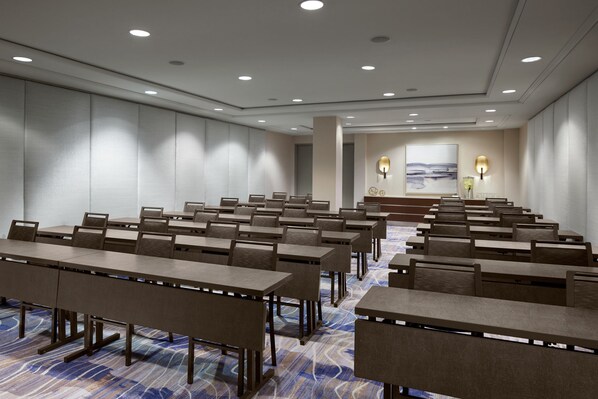 A conference room with rows of tables and chairs, recessed ceiling lights, and a large painting on the back wall.