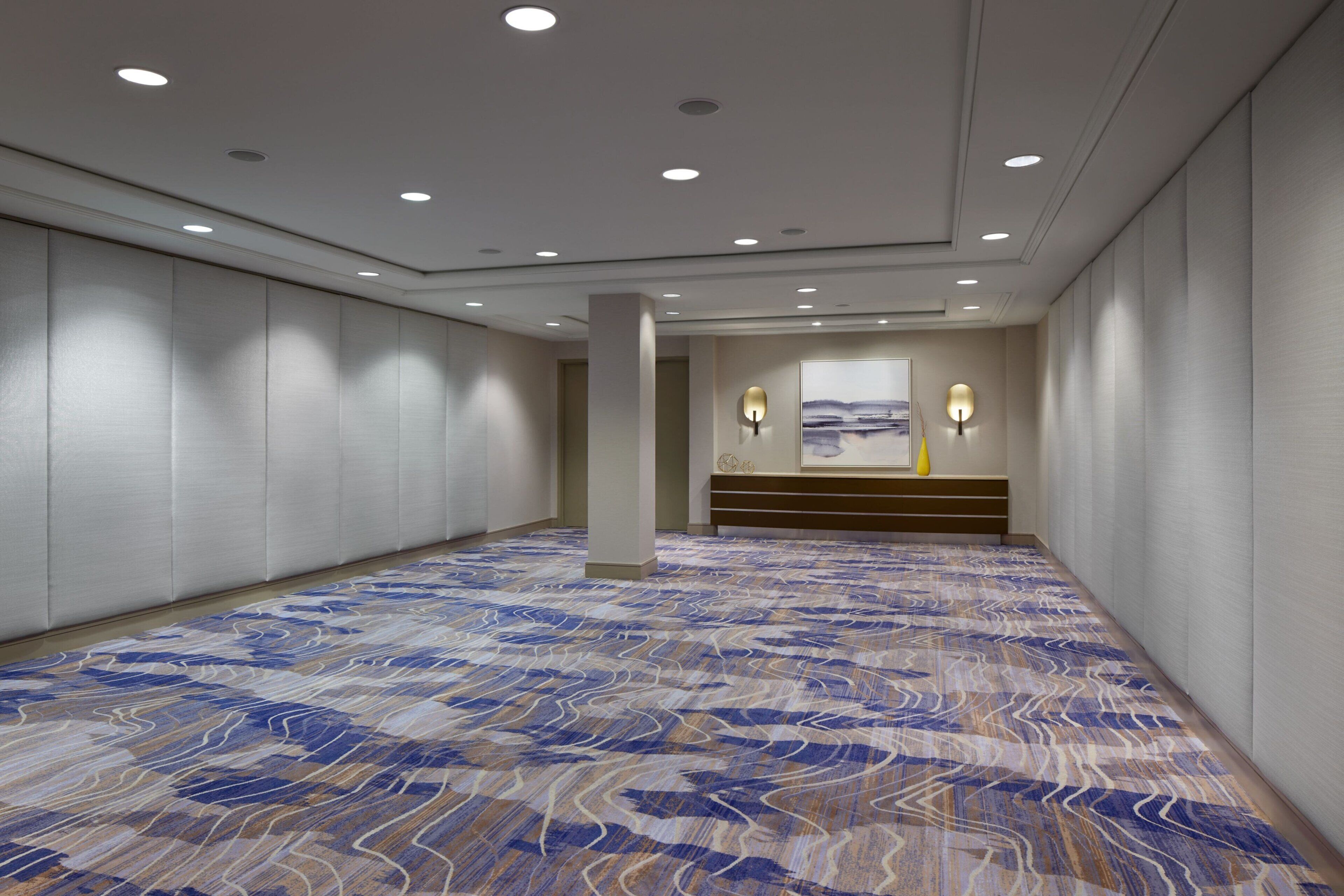 Um hall de hotel espaçoso com carpete estampado, iluminação embutida e um banco com luminárias fixadas na parede.