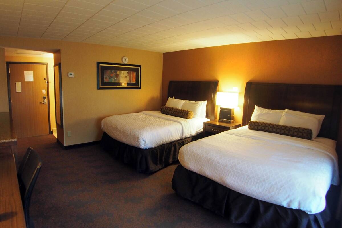 Ein Hotelzimmer mit zwei Betten, einem Schreibtisch und einem Stuhl. An der Wand hängt ein gerahmtes Bild und auf dem Schreibtisch steht eine Lampe.