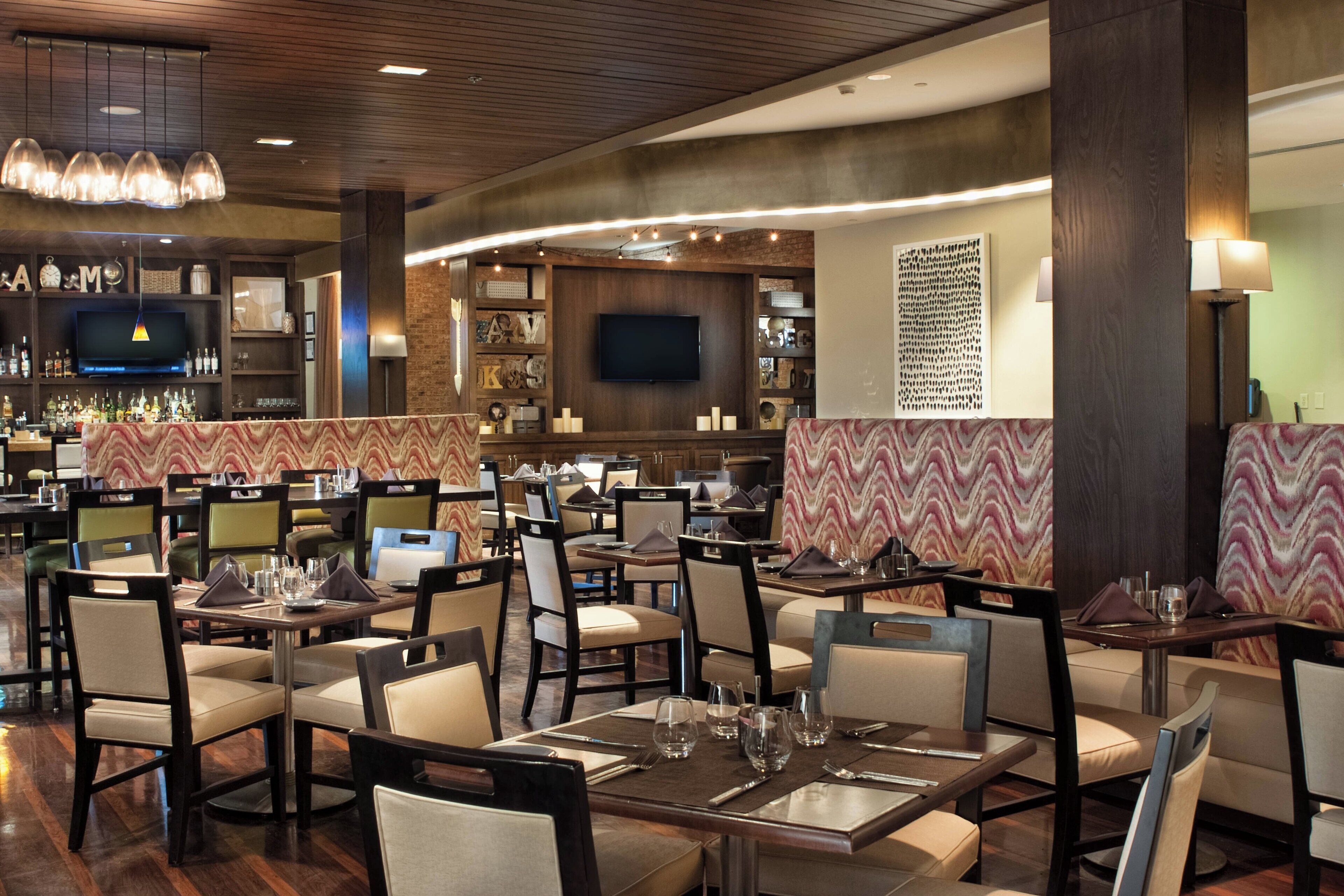 A restaurant with multiple dining tables and chairs, a bar area with a television, and a wall-mounted artwork.