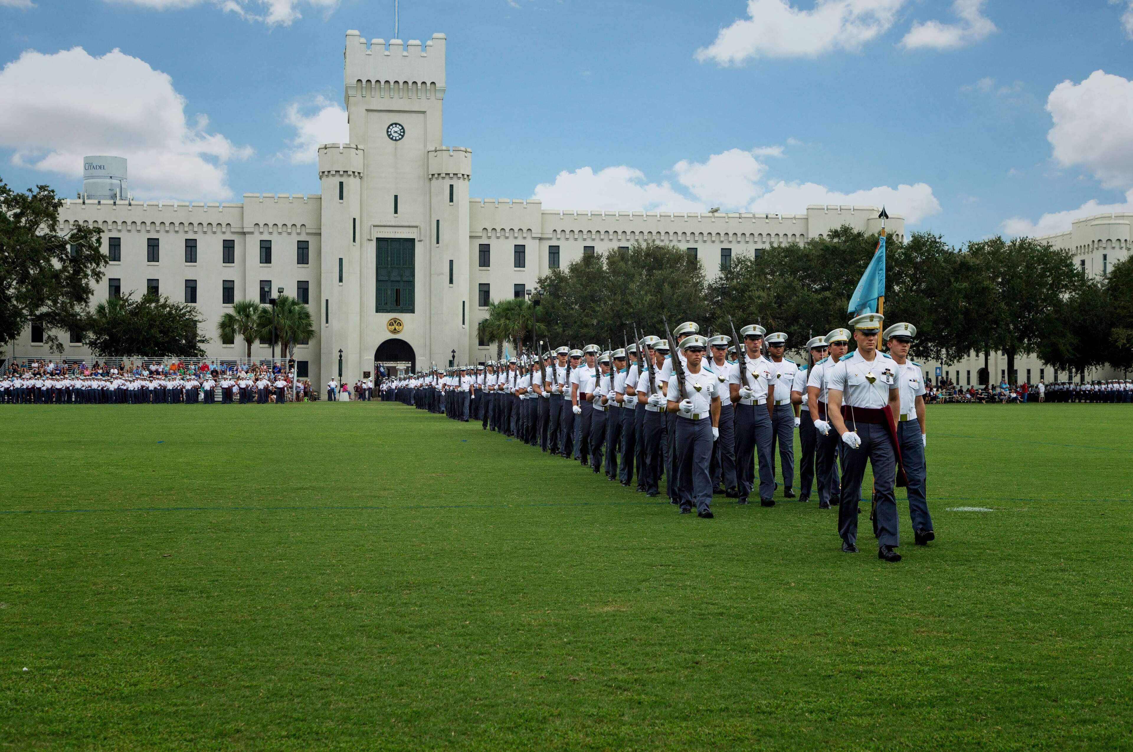 A formal military ceremony with personnel in uniform standing in formation on a grassy field in front of a large building with a clock tower.