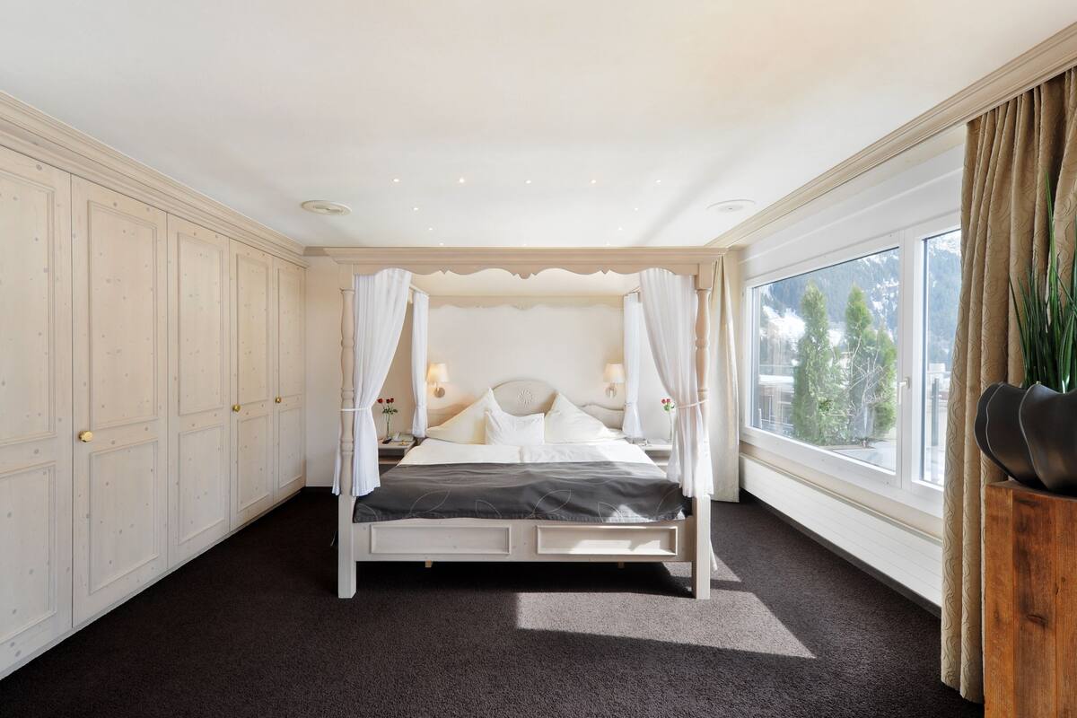 A bedroom with a four-poster bed, a large window with a view of trees, and a wooden nightstand with a lamp.