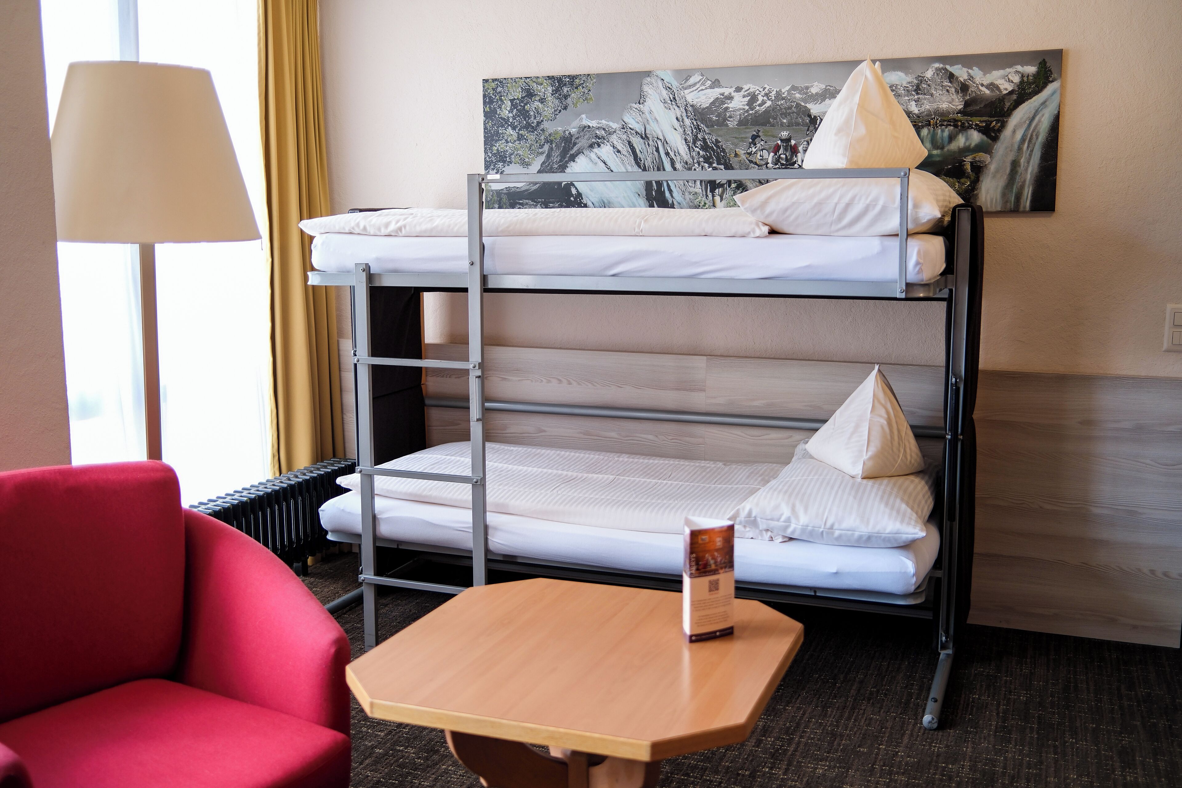 A bunk bed with a view of a mountain landscape, a red chair, and a table with a brochure.