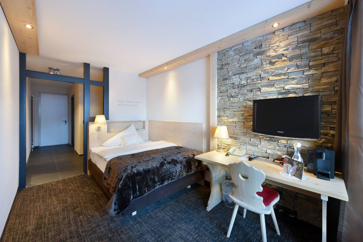 A hotel room with a bed, desk, chair, television, and stone wall.