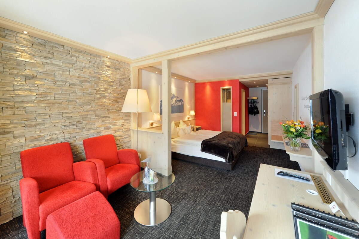 A hotel room with a stone wall, a bed, a red sofa, a glass table, and a TV.