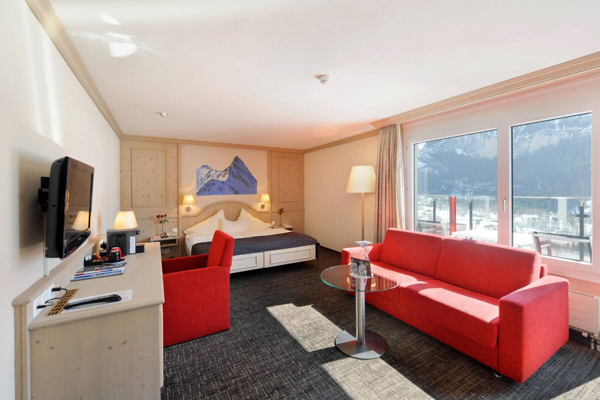 A hotel room with a bed, a red sofa, a glass coffee table, a desk with a TV, and a view of mountains through large windows.