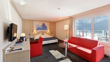 A hotel room with a bed, a red sofa, a glass coffee table, a desk with a TV, and a view of mountains through large windows.