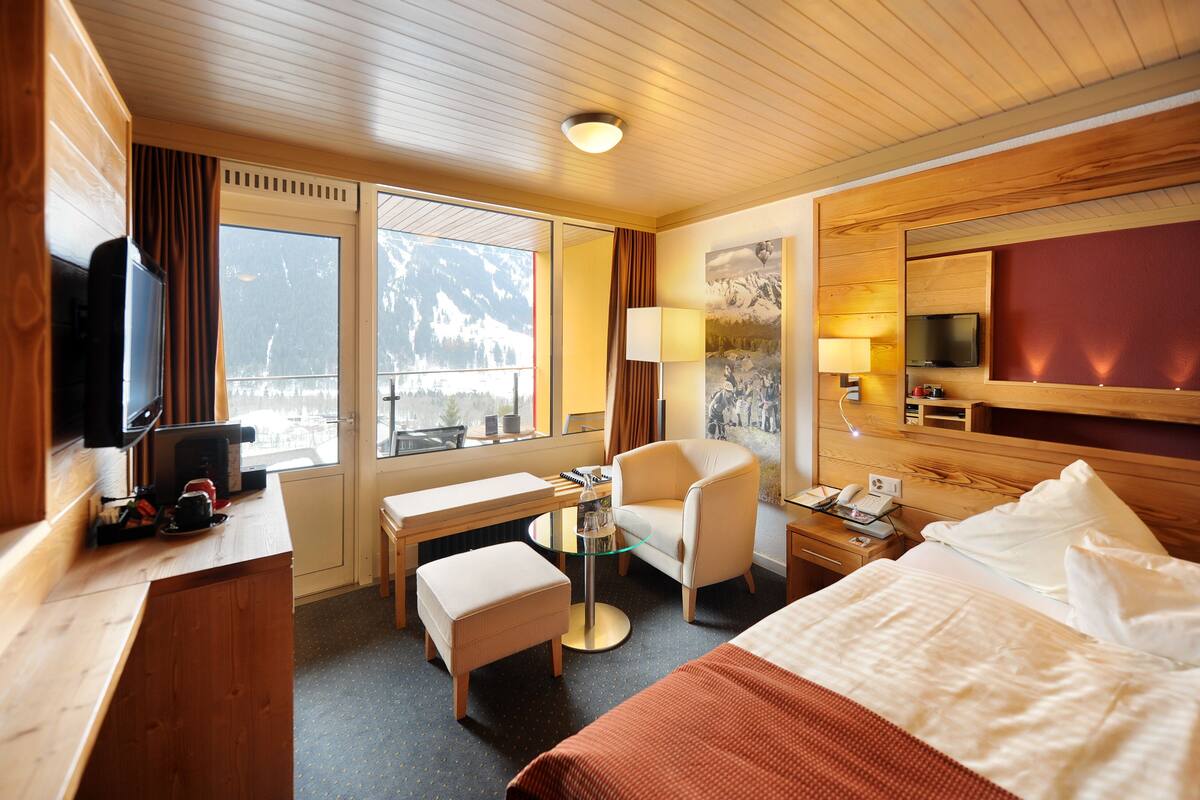 A hotel room with a wooden ceiling, a bed, a desk with a chair, a television, and a window with a mountain view.
