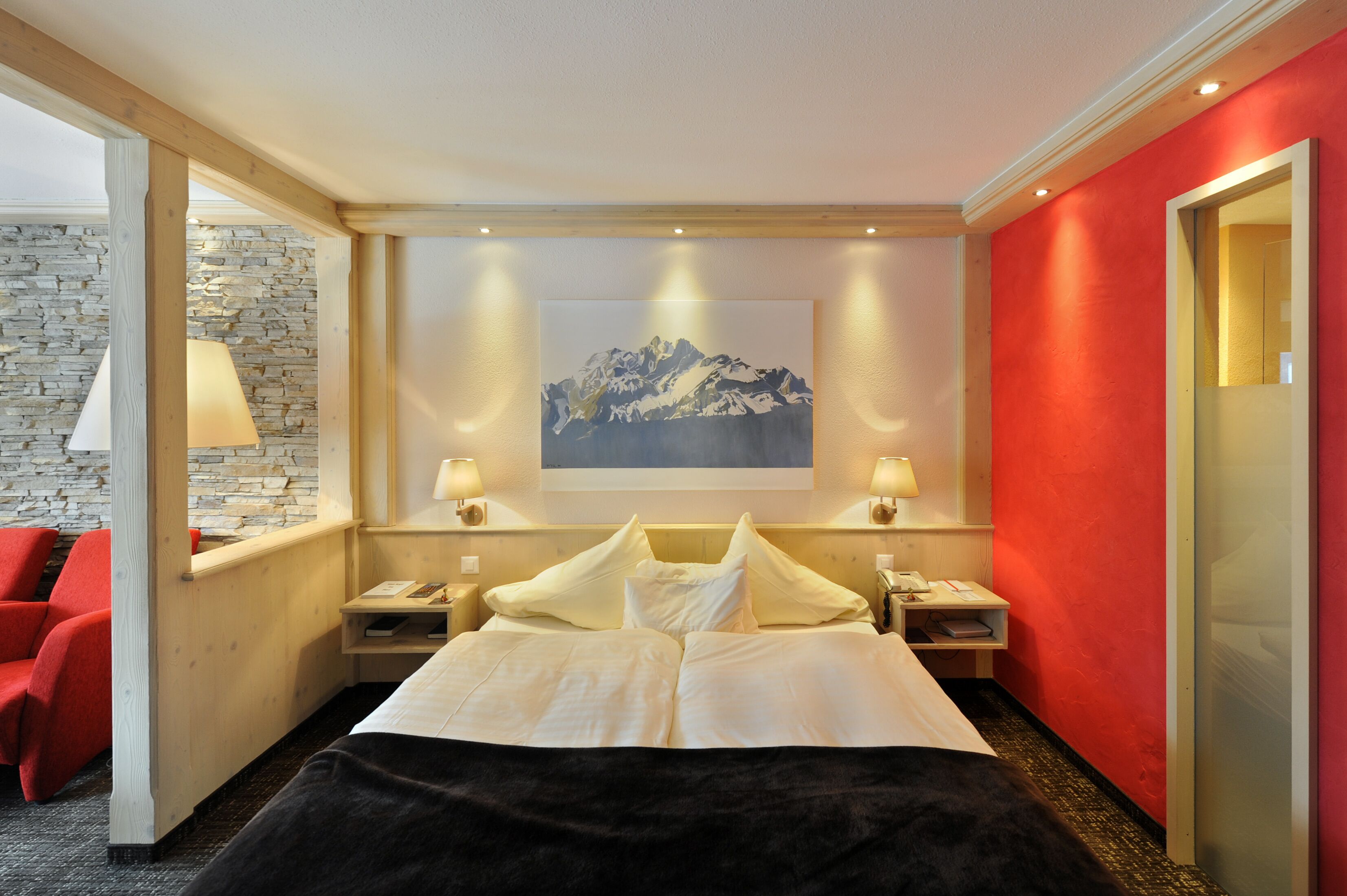 A hotel room with a bed, a nightstand, a lamp, a red wall, and a mountain painting on the wall.