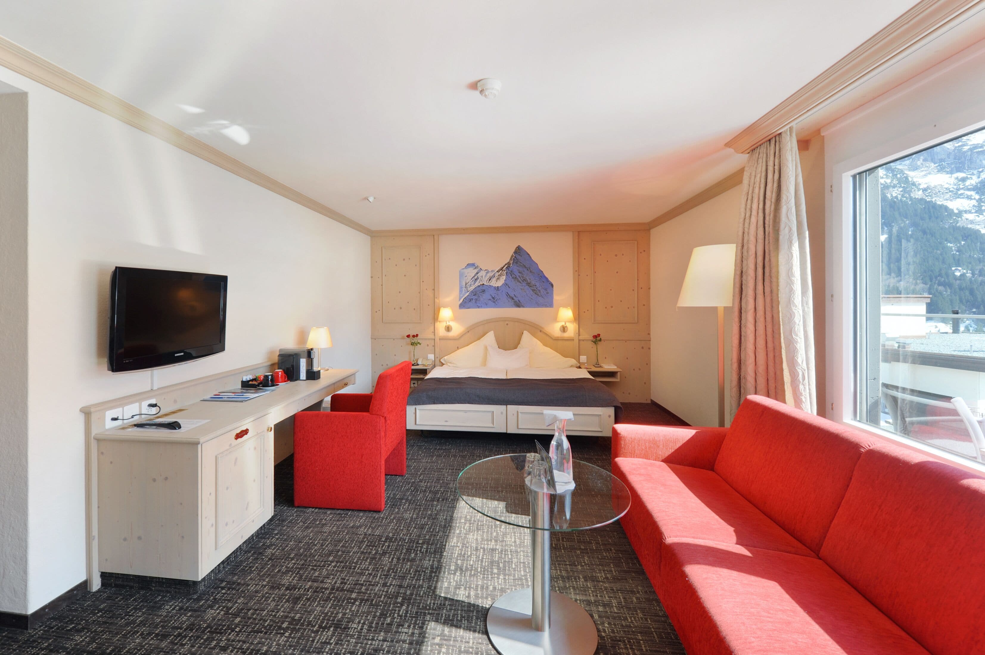 A hotel room with a bed, a desk, a television, a red sofa, a glass coffee table, and a view of the mountains.