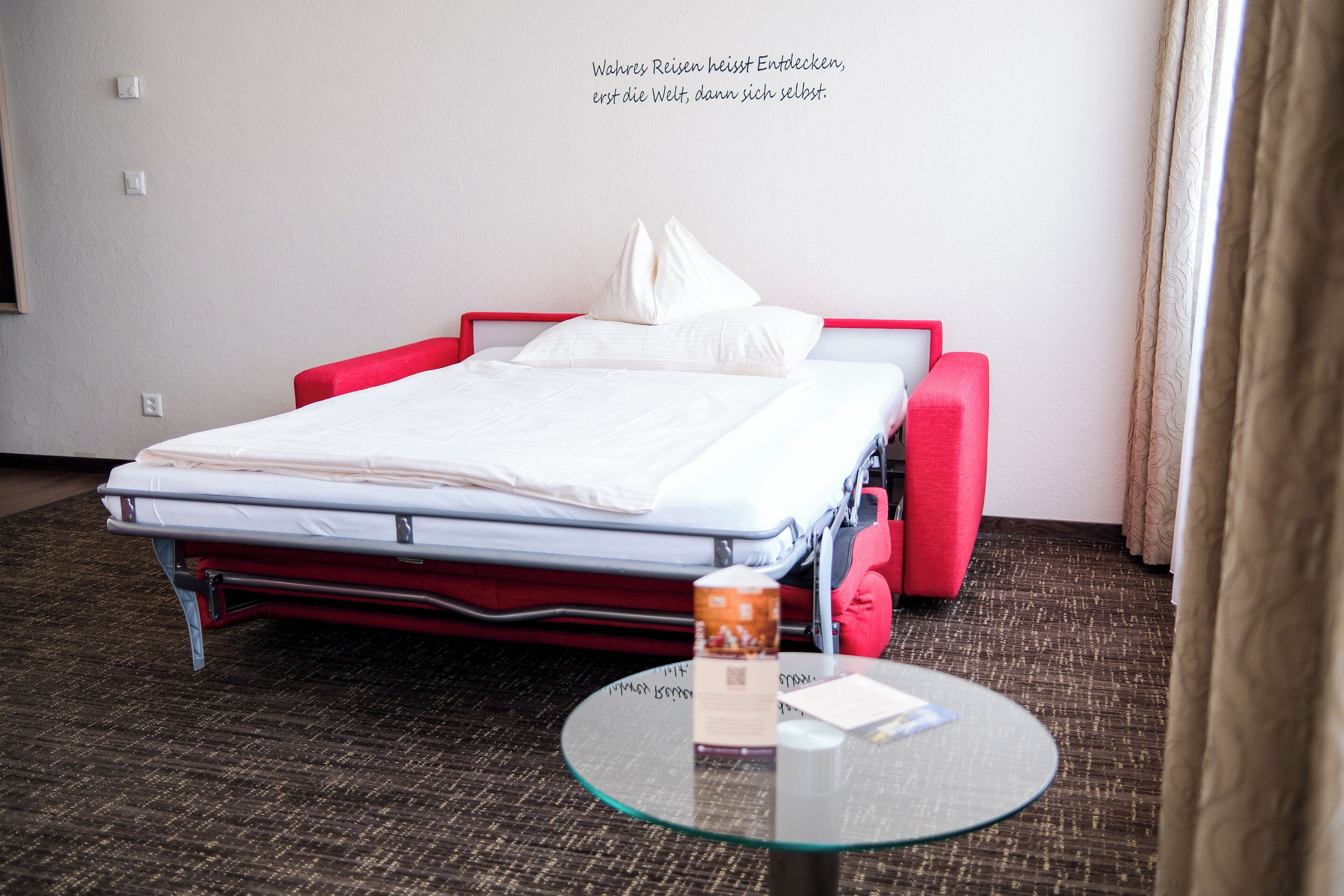 A hotel room with a single bed, a red chair, a small table with a glass top, and a wall with a quote in German.