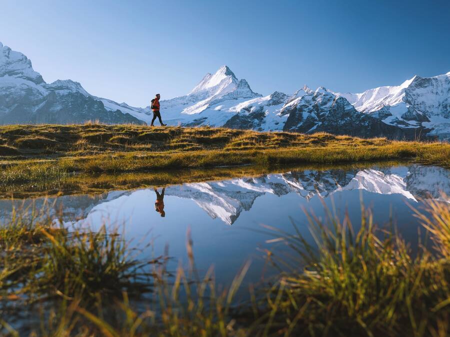 A person hiking in a mountainous area with snow-capped peaks in the background and a reflective water body in the foreground.