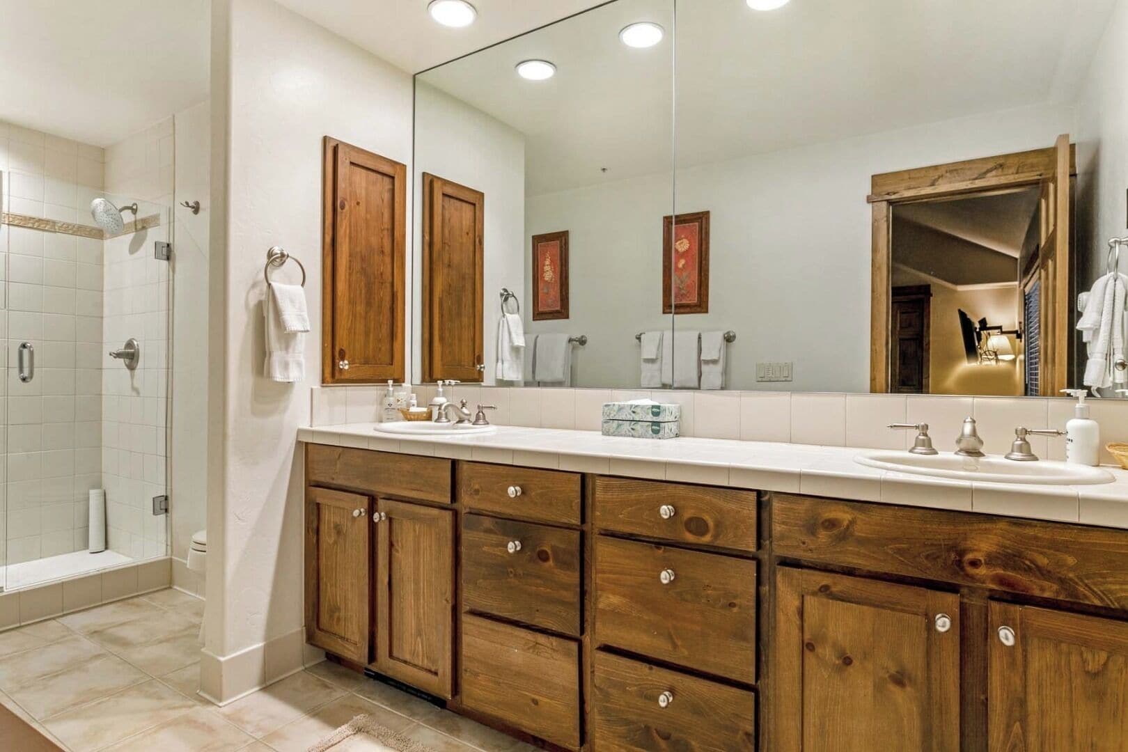Un baño moderno con un espejo grande, armarios de madera, zona de ducha y encimera blanca.