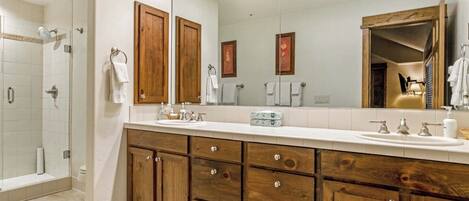 Un baño moderno con un espejo grande, armarios de madera, zona de ducha y encimera blanca.