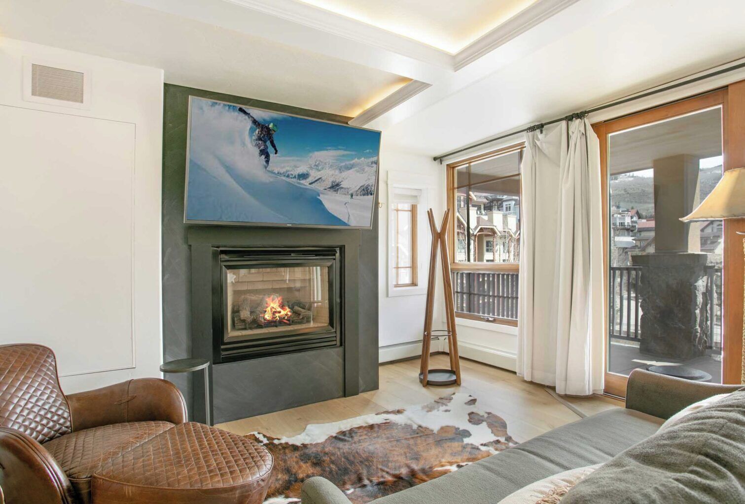 A living room with a fireplace, a large painting of a snowy mountain, leather furniture, and a view of a balcony with a railing.