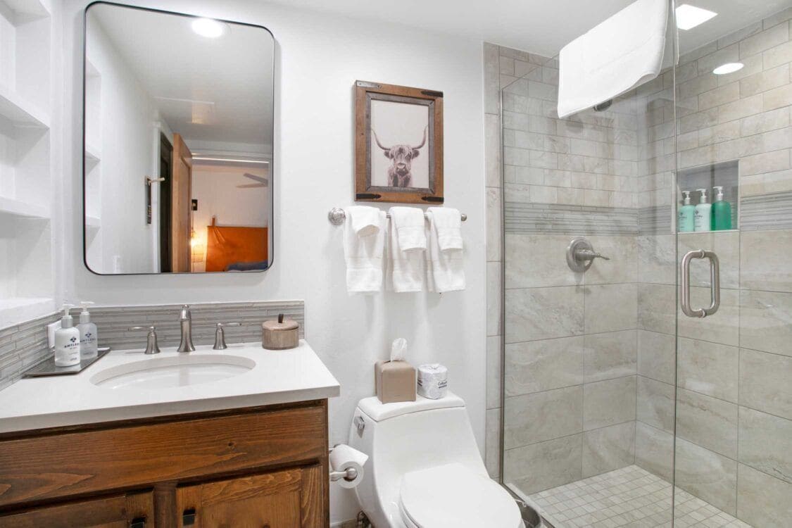 A modern bathroom with a large mirror, a sink with toiletries, a toilet, and a glass-enclosed shower.